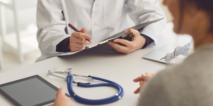  Medical professional writing on a clipboard while sitting at a table that has a stethoscope, digital tablet, and computer on it