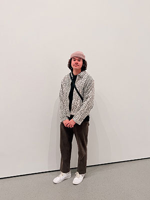 Antoine has long, curly black hair under a beanie. He's standing with hands clasped in a white leopard print jacket.