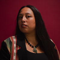 A woman with long dark straight hair looks seriously at the camera against a red background