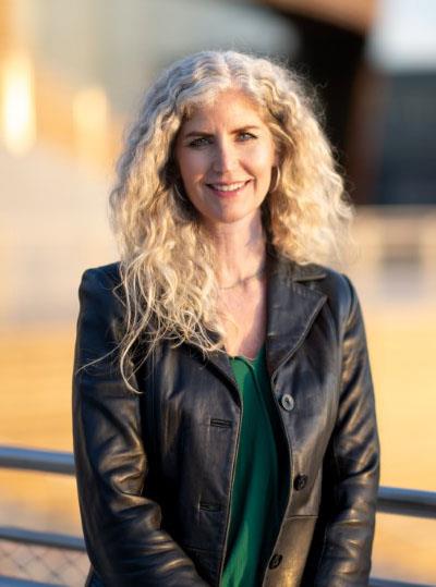 Angela Brady is smiling.  She has long, white-blond curly hair and a black leather jacket. 