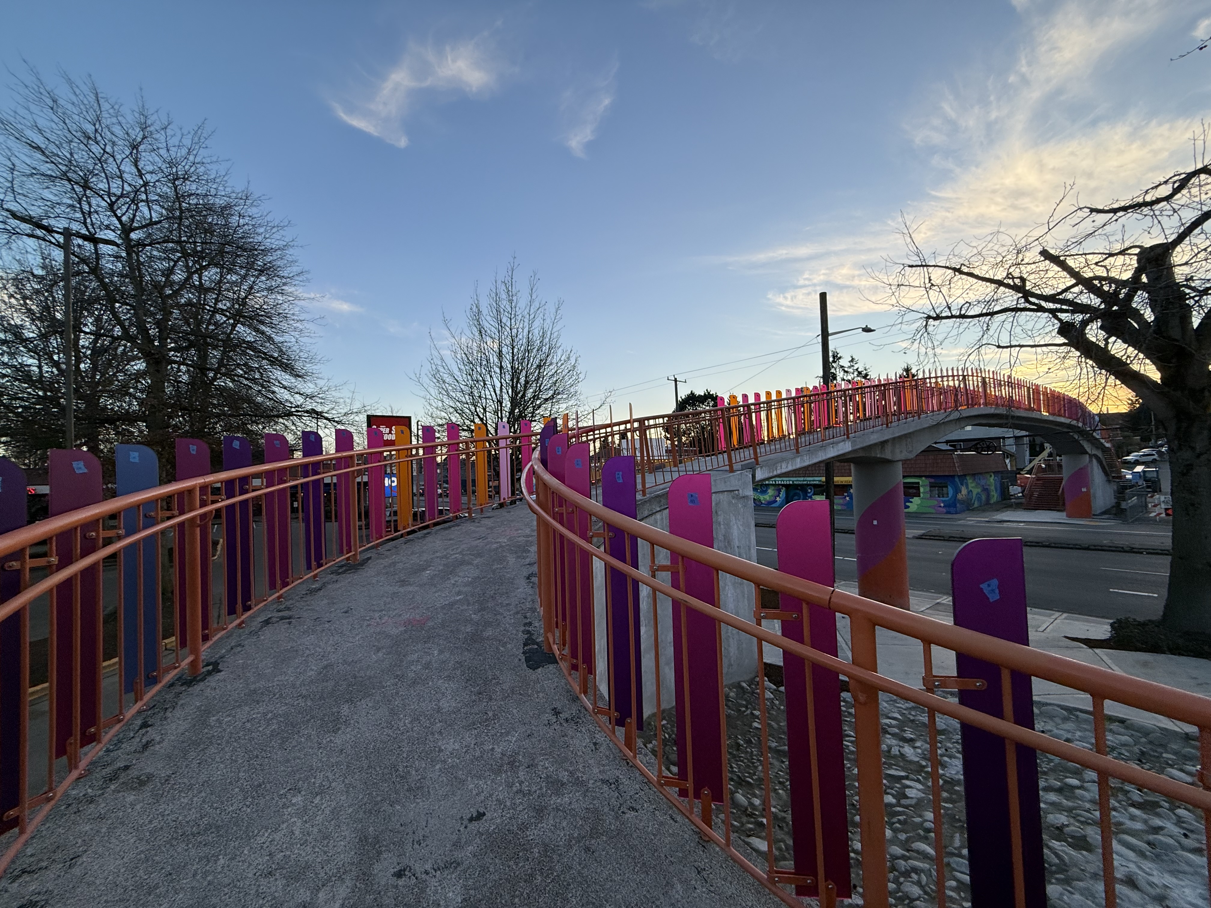 A pedestrian bridge with orange railings and pink-purple panels curves over Aurora Ave N at sunset. Trees and a mural-covered building are in the background.