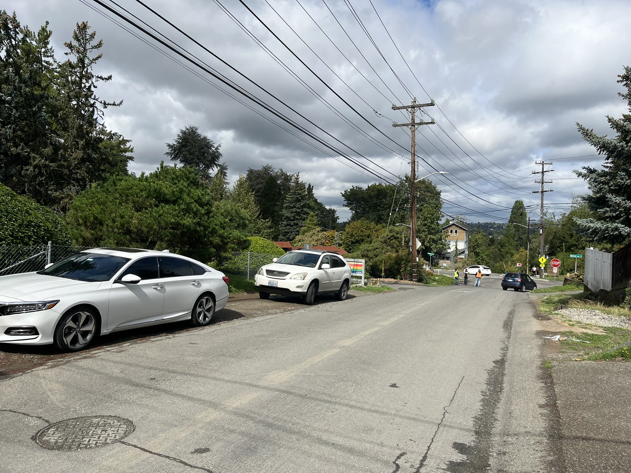 A street view of 55th Ave S Sidewalk, facing north, with cars parked on the west side of the road, utility poles and wires overhead, and pedestrians visible in the distance near an intersection without sidewalks.
