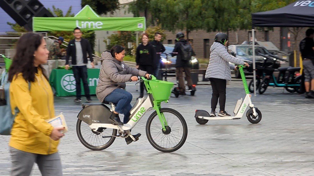 People riding a Lime bike and a Lime scooter at a Bike & Scoot Demo Day in Capitol Hill. Organization tents are in the background. 