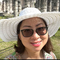 Linda is wearing a large sunhat, sunglasses, and is smiling.  She has dark shoulder-length hair.