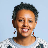 Tsega has short black and gray hair, and is wearing earrings and a sweater.