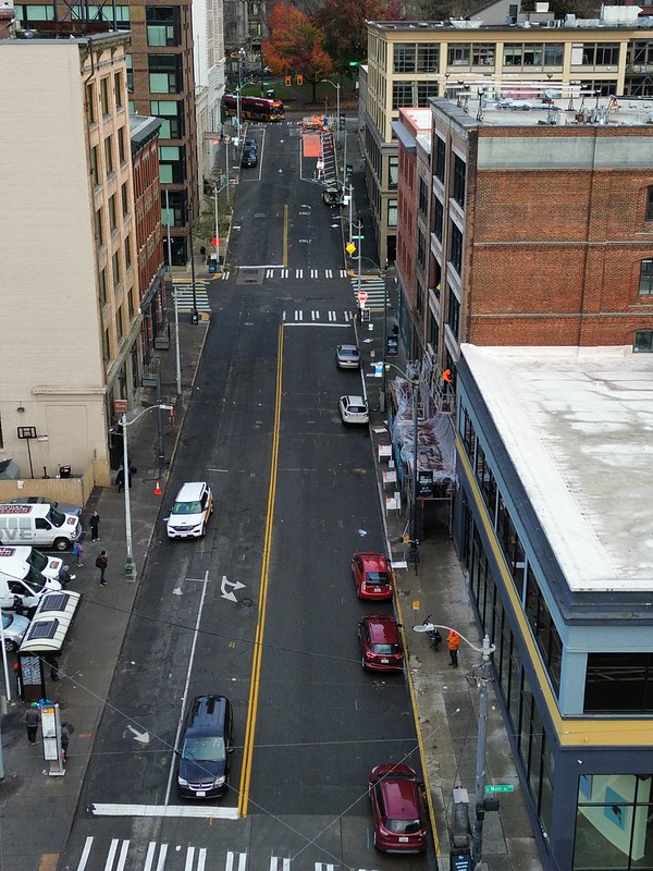 A photo taken with a drone, showing 3rd Ave facing north from S Main St to Yesler Way. Cars are parked on the east side of the street and vehicles travel southbound down the patchy, bumpy street &nbsp;