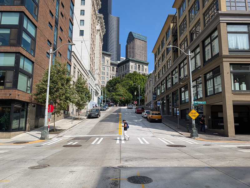 A person walking in the empty intersection at 3rd Ave and S Washington. Cars are parked on either side of the street in the background
