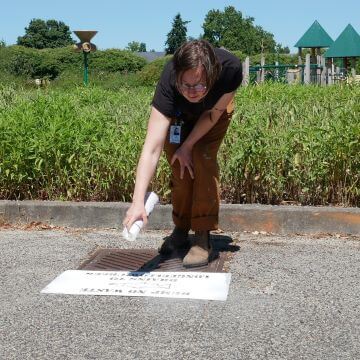A person stands on a grate, using one arm to spray paint a stencil.