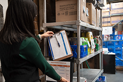 A grocer checks information on a clipboard against an inventory list on a shelf full of items in a grocery warehouse.
