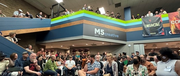 A crowd of people are seated inside the M5 Creative Building waiting for a presentation to begin.