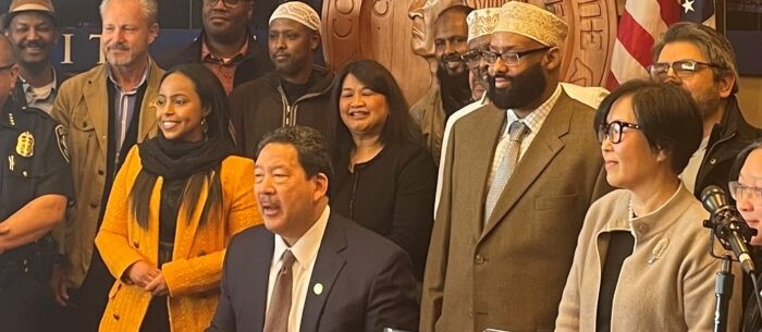 Mayor Bruce Harrell sits at table in front of City of Seattle seal and several people standing behind him at City Hall.