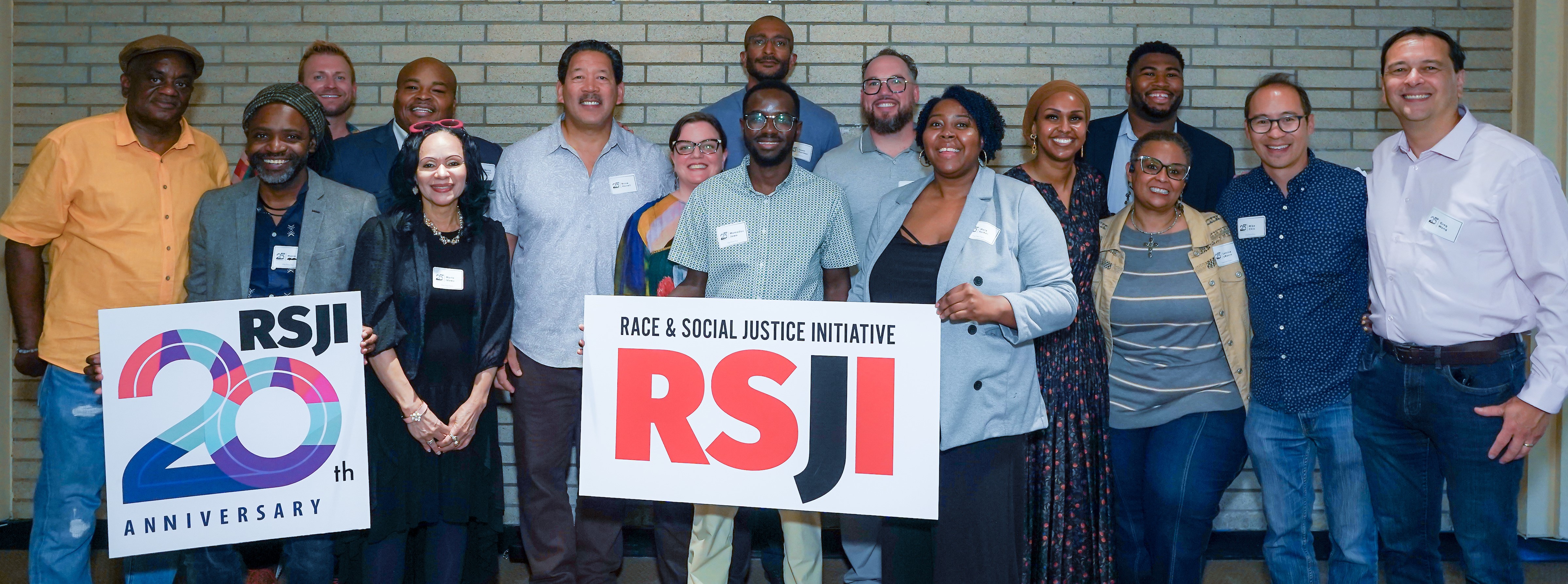 Seattle Office for Civil Rights staff, Seattle Mayor Bruce Harrell, and Deputy Mayor Greg Wong stand for a photo at the RSJI 20th Anniversary Reception in August 2024. On the left, people are holding a sign: "20th Anniversary)". On the right is another held sign with the RSJI logo.