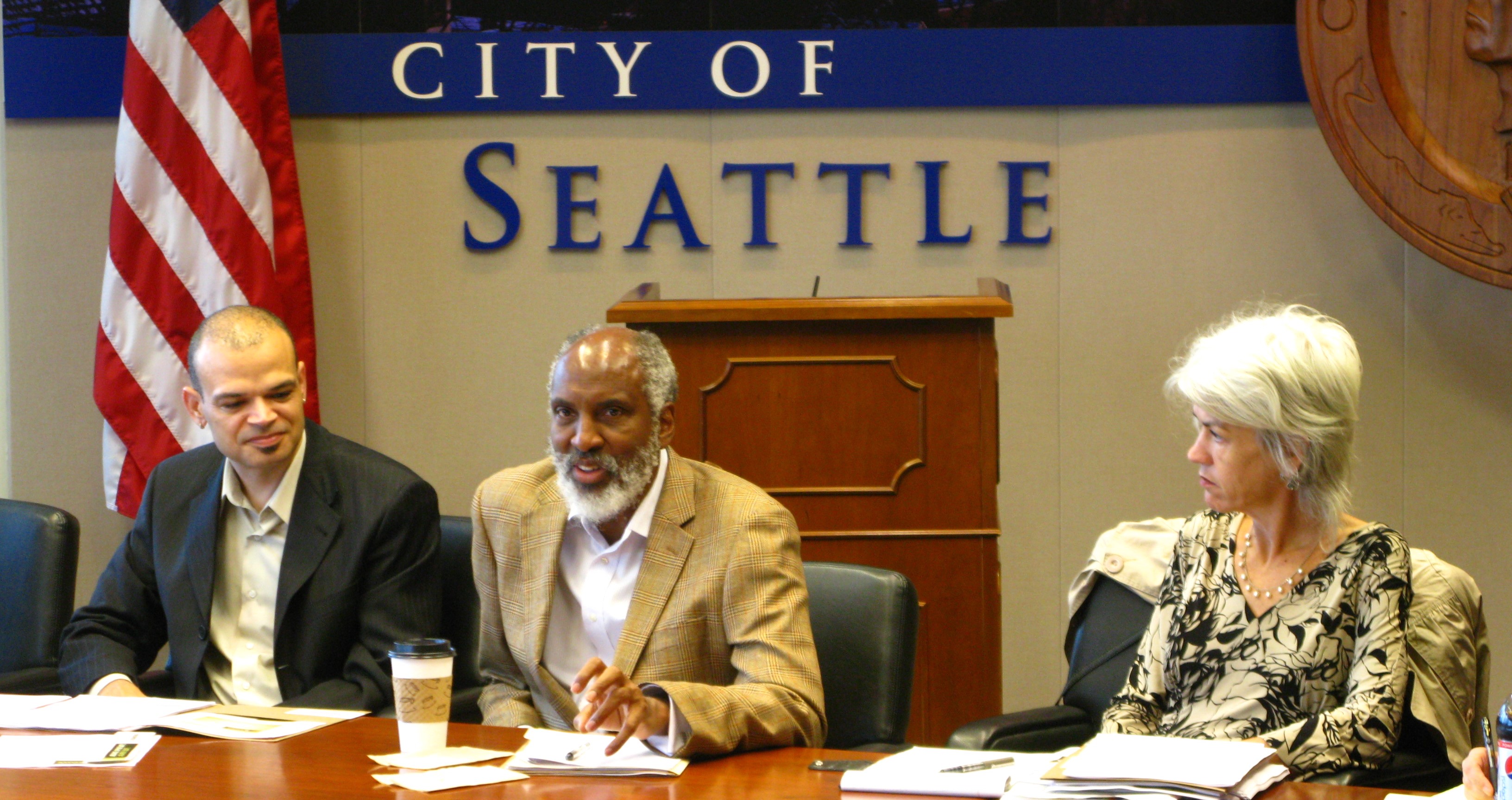 Former RSJI Manager Glenn Harris (left) and former Seattle Office for Civil Rights Director Julie Nelson (right) sit with john a. powell (middle) during an RSJI Summit event in 2011. A "City of Seattle" sign is in the background.