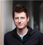 Portrait of Dylan Glosecki smiling at the camera wearing a dark button up shirt in front of a grey background.