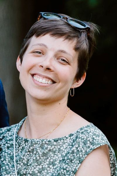 Portrait of Xio smiling broadly at the camera in a floral print top in front of a dark background.
