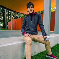 Model Cody Volk sitting and wearing a gray jacket with a maroon collar and tan plaid trousers.