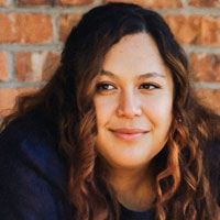 Jesse has long, dark, wavy hair and is smiling in front of a brick background.