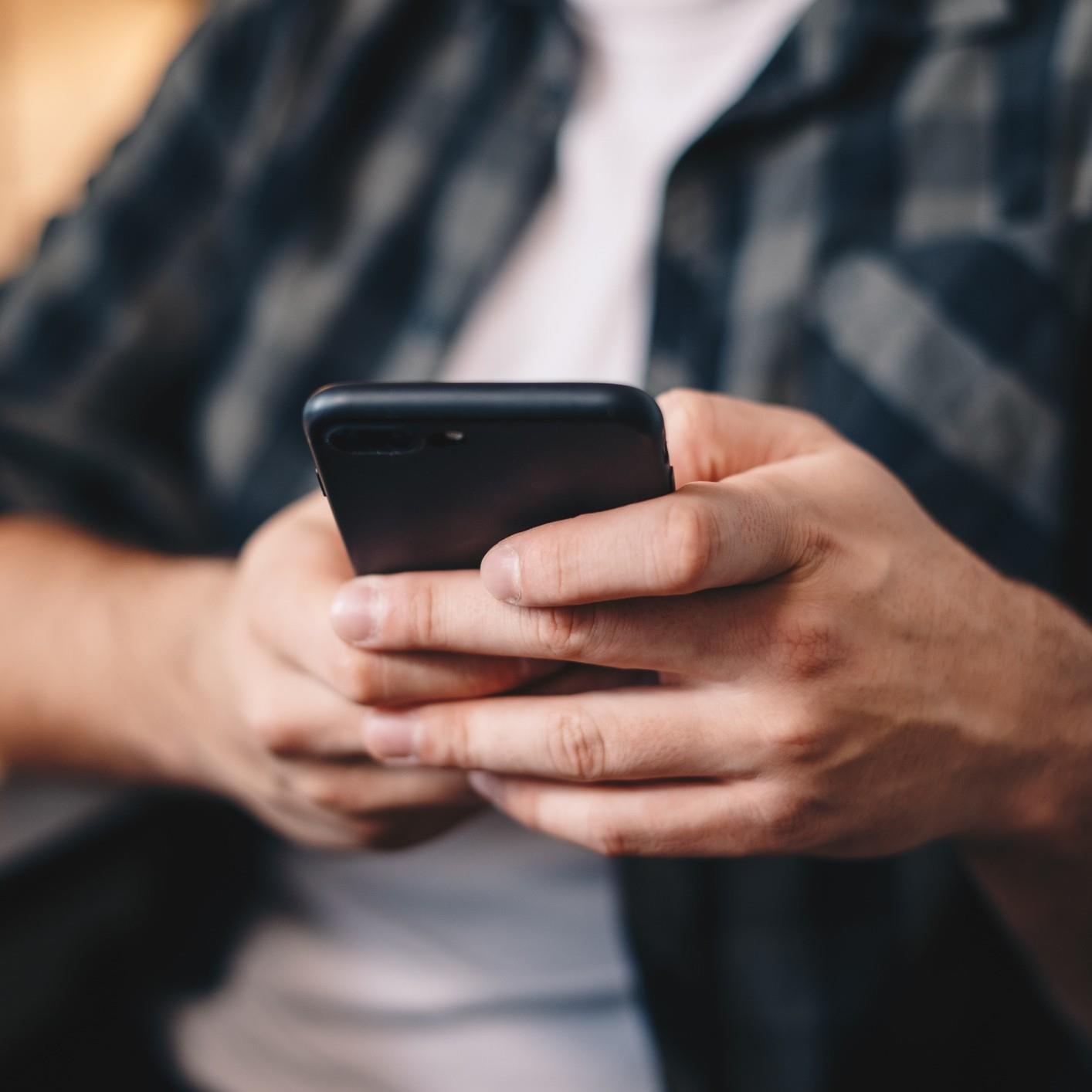 A person in a grey and black plaid flannel button down shirt with a white shirt underneath is holding a mobile phone device in both hands. The focus of the image is on the mobile device.