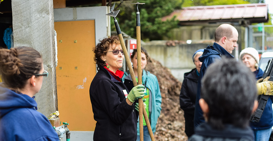 A community gardener holding up two garden tools and talking to a group of people.