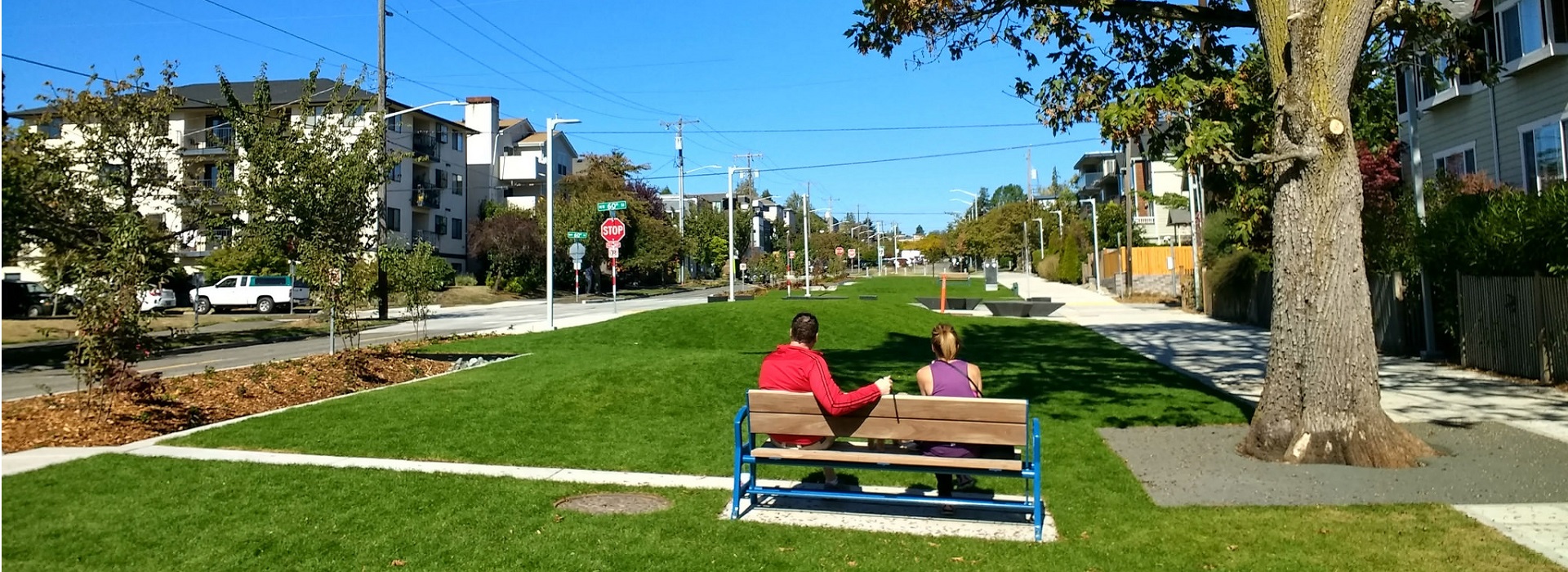 Two people sit on a bench at one end of a narrow park in a residential neighborhood.