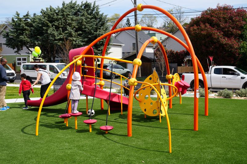 Young children play on a colorful, modern playground structure with bright red, orange, and yellow elements. Adults supervise nearby, and balloons and parked vehicles add to the festive neighborhood atmosphere.