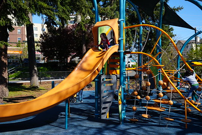 A young child wearing a pink shirt sits at the top of a tall, curved orange slide on a blue-and-orange playground surrounded by trees.