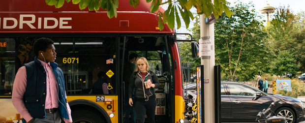 A woman exits a Rapid Ride bus while a man walks by on the sidewalk.  The Spaceneedle is in the distance. 