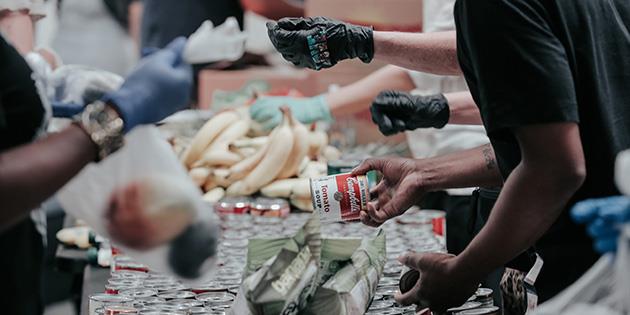 Overhead view of an individual sitting on a wooden floor, surrounded by papers and holding a document. The person is wearing a striped shirt and ripped blue jeans. The focus is on the document in their hands, which appears to be a bill or statement.