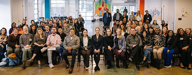 Mayor Katie B. Wilson's transition team and committee, a diverse group of people pose together indoors in a bright community space, seated and standing in rows, smiling toward the camera.