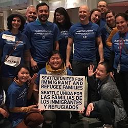 A multiracial group of smiling Seattle staff and volunteers with a sign that reads: SEATTLE UNITED FOR IMMIGRANT AND REFUGEE FAMILIES | SEATTLE UNIDA POR LAS FAMILIAS DE LOS INMIGRANTES Y REFUGIADOS.