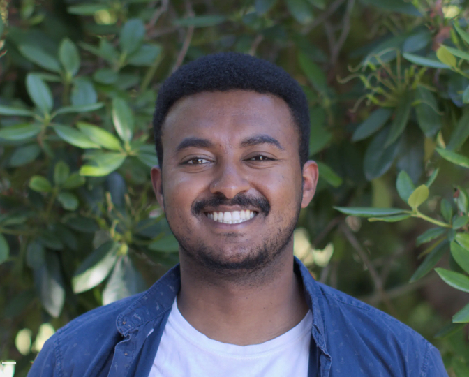 Portrait of Kelabe smiling broadly at the camera in a blue button up shirt in front of a background of green leaves.